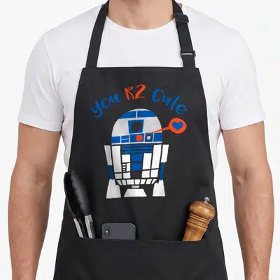 Cool valentine's day apron - you R2 Cute text arching over cartoon R2-D2 with blue heart in orange bubble — shown on a black cotton canvas apron against a white background