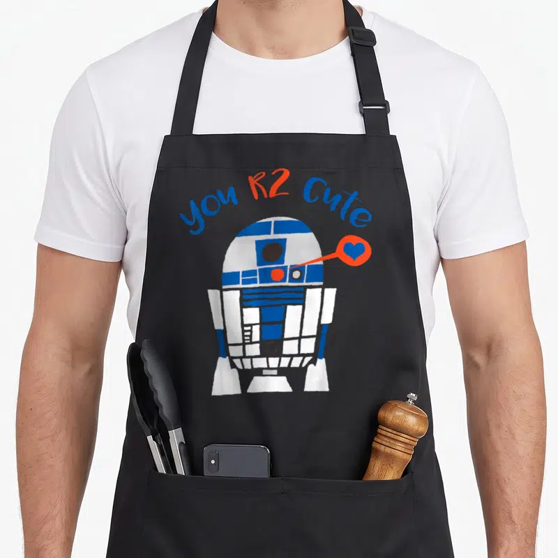 Cool valentine's day apron - you R2 Cute text arching over cartoon R2-D2 with blue heart in orange bubble — shown on a black cotton canvas apron against a white background