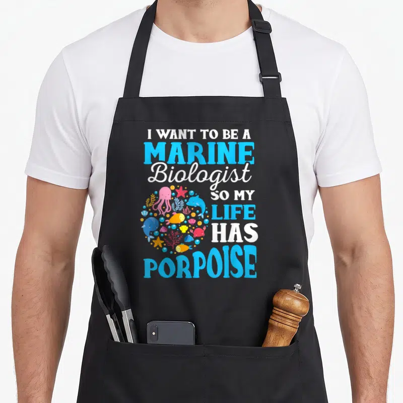 Biology marine biologist apron - I WANT TO BE A MARINE Biologist SO MY LIFE HAS PORPOISE text with circular sea — shown on a black cotton canvas apron against a white background