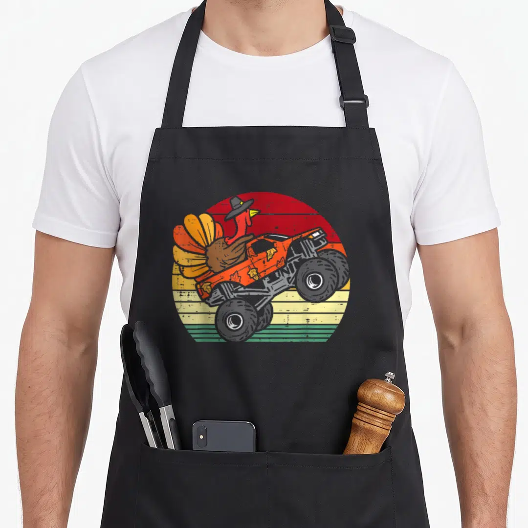 Turkey truck aprons - Cartoon turkey in hat driving orange monster truck on striped retro background — shown on a black cotton canvas apron against a white background