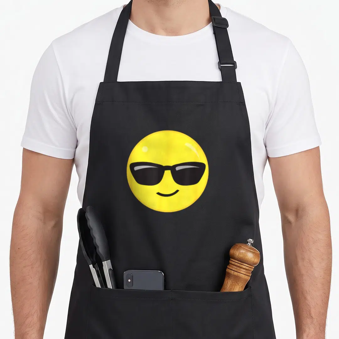 Sunglass face on apron - Yellow cartoon smiley face wearing black sunglasses printed on apron front — shown on a black cotton canvas apron against a white background