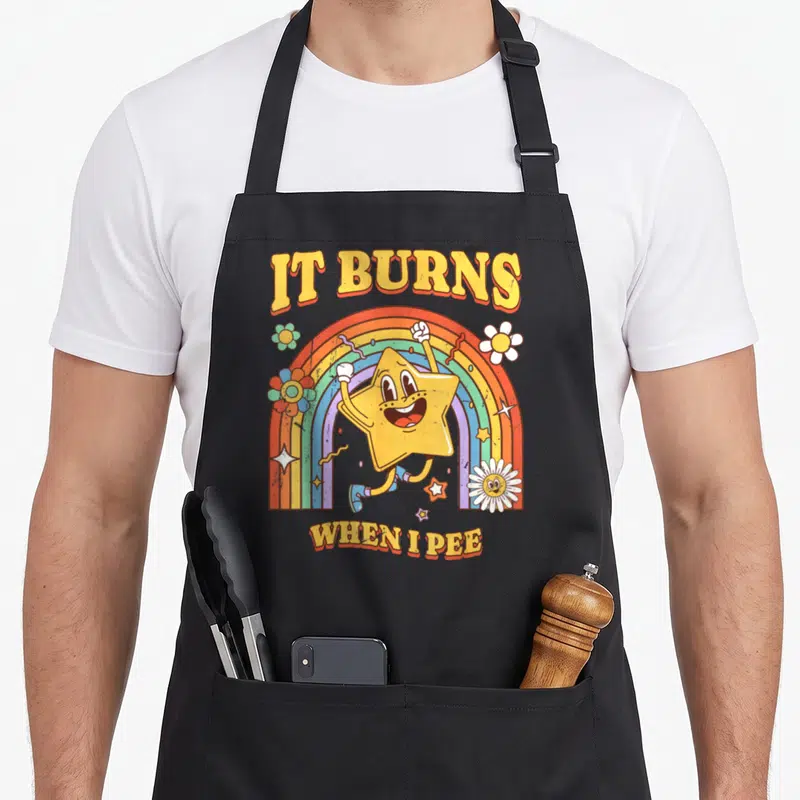 Burn sarcastic apron - IT BURNS WHEN I PEE text with yellow star and rainbow illustration — shown on a black cotton canvas apron against a white background