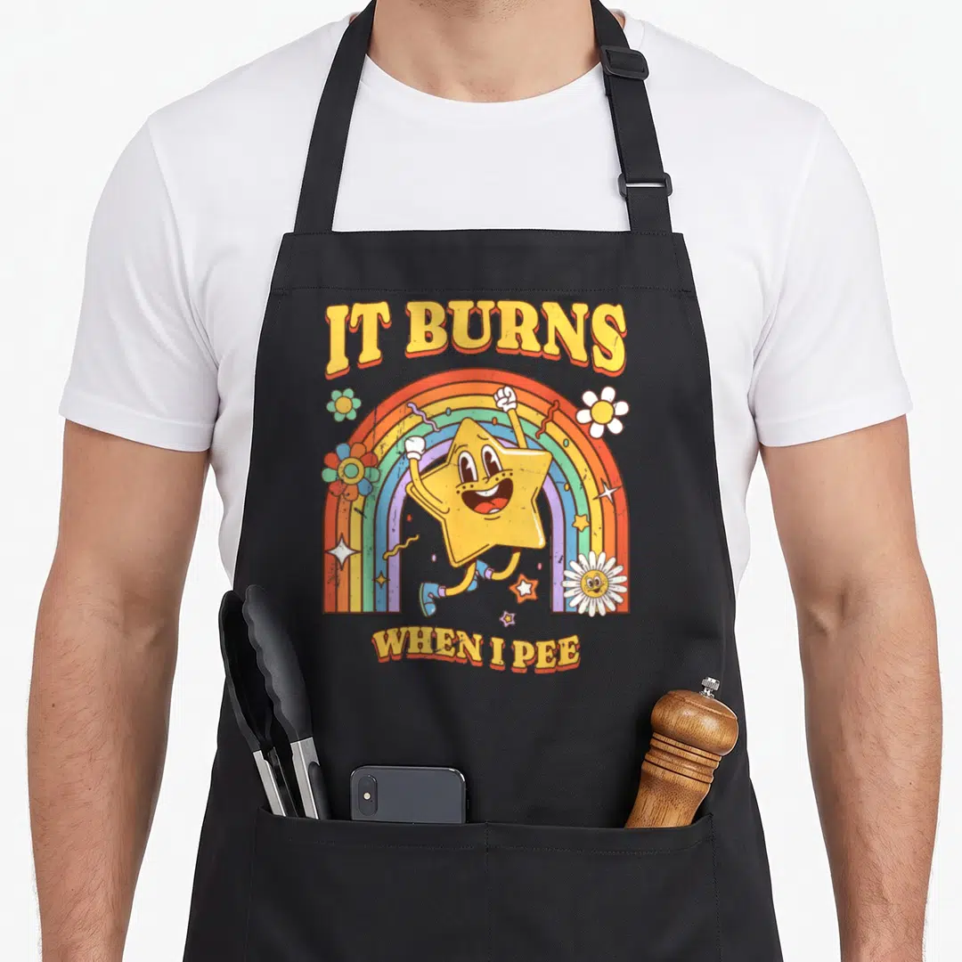 Burn sarcastic apron - IT BURNS WHEN I PEE text with yellow star and rainbow illustration — shown on a black cotton canvas apron against a white background