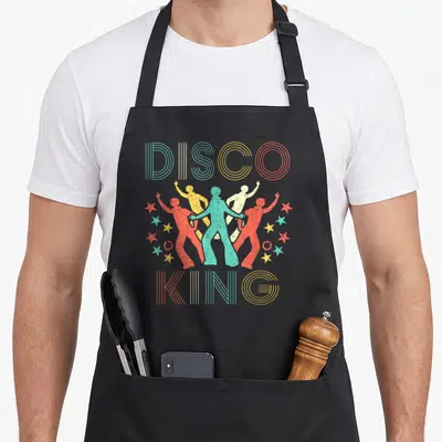 Dance king apron - Four retro disco dancers in teal yellow red and white flanked by stars and striped text — shown on a black cotton canvas apron against a white background