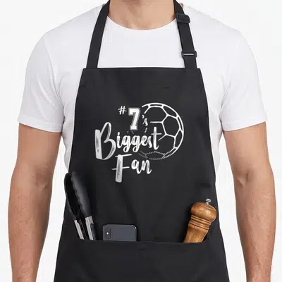 Number family aprons - #7's Biggest Fan text and white soccer ball outline on black fabric — shown on a black cotton canvas apron against a white background
