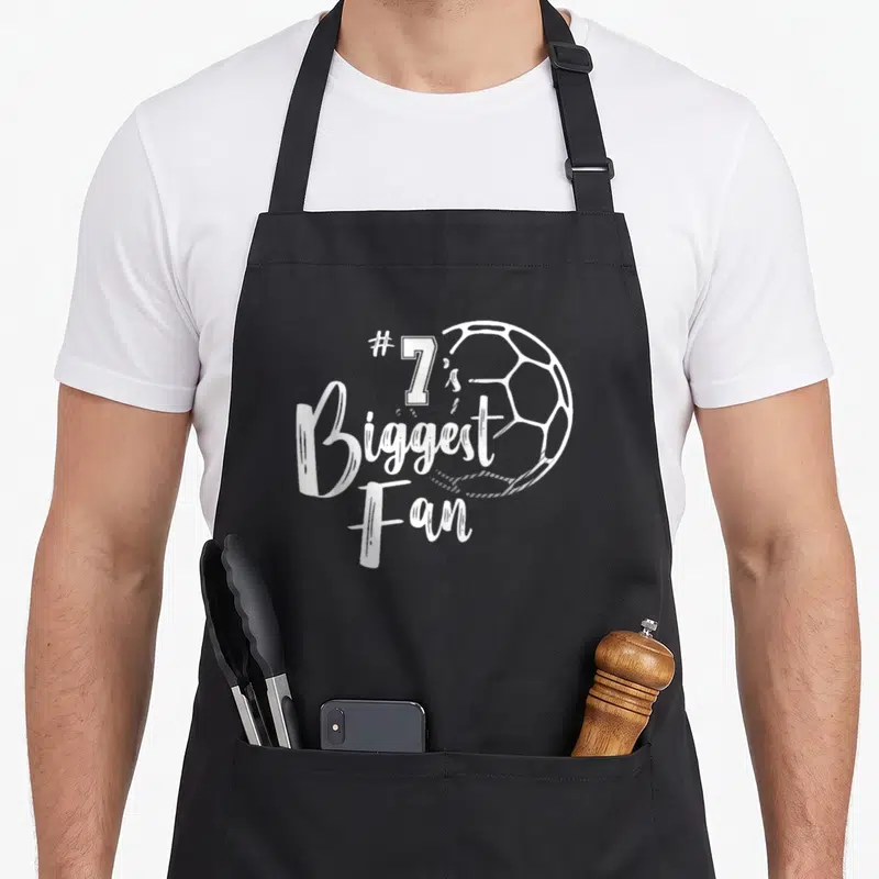 Number family aprons - #7's Biggest Fan text and white soccer ball outline on black fabric — shown on a black cotton canvas apron against a white background