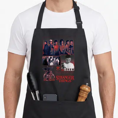 Eleven stranger apron - Collage of Eleven characters, Upside Down text, and Demogorgon illustration — shown on a black cotton canvas apron against a white background