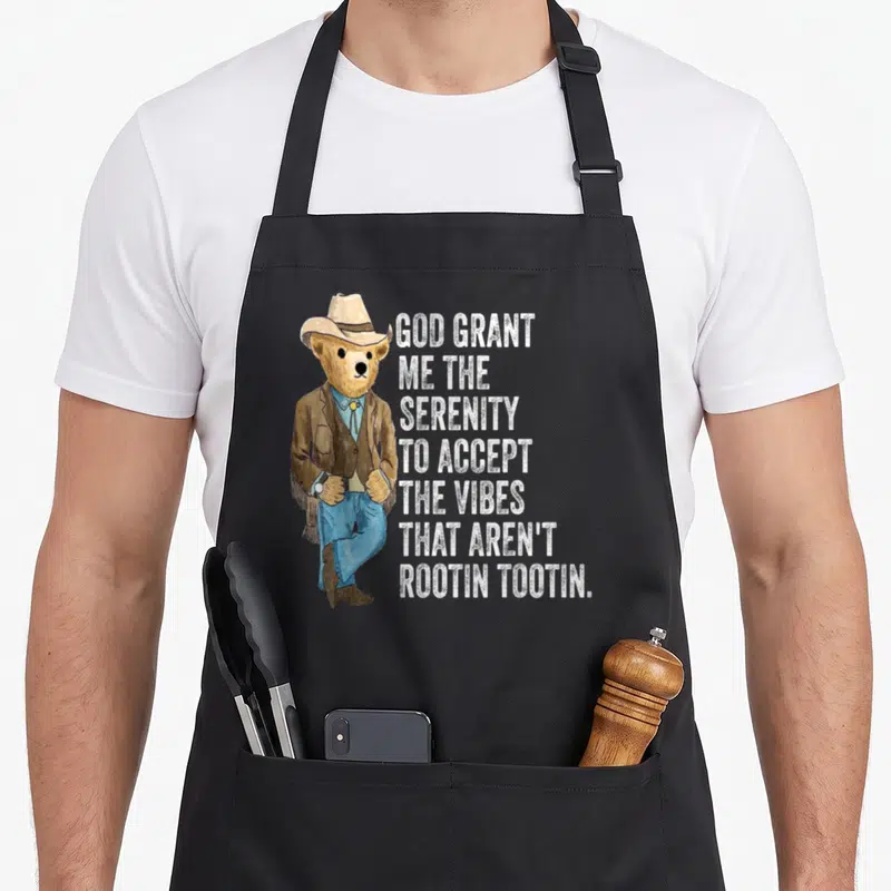 Grant serenity apron - Cowboy bear in brown jacket and blue jeans next to seven lines of white text about vibes — shown on a black cotton canvas apron against a white background