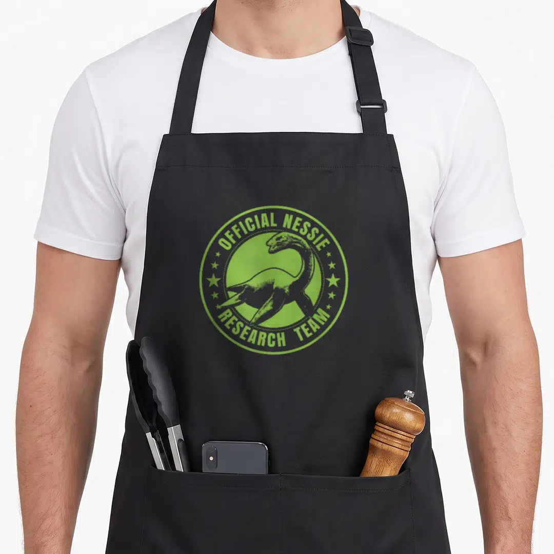 Ness scottish boy apron - Lime green circular Nessie emblem with text Official Nessie Research Team and stars — shown on a black cotton canvas apron against a white background