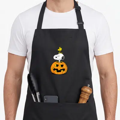 Woodstock halloween apron - Snoopy emerging from an orange jack-o-lantern with Woodstock on his head in white — shown on a black cotton canvas apron against a white background