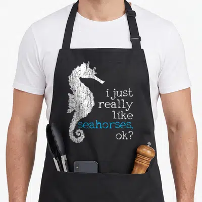 Seahorse fish apron - Distressed white seahorse silhouette beside stacked text i just really like seahorses ok? — shown on a black cotton canvas apron against a white background