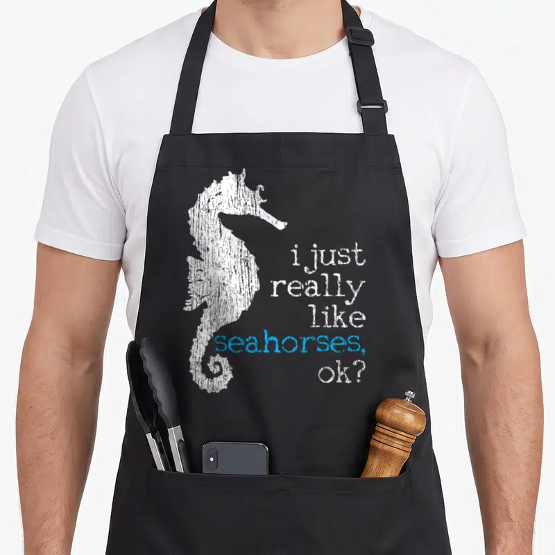 Seahorse fish apron - Distressed white seahorse silhouette beside stacked text i just really like seahorses ok? — shown on a black cotton canvas apron against a white background