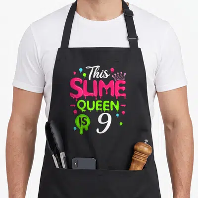 Slime birthday queen apron - Text This SLIME QUEEN IS 9 with pink dripping letters green blob and crown — shown on a black cotton canvas apron against a white background