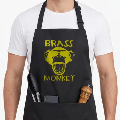 Brass trending graphic and apron - Yellow cartoon monkey head between BRASS above and MONKEY below text — shown on a black cotton canvas apron against a white background