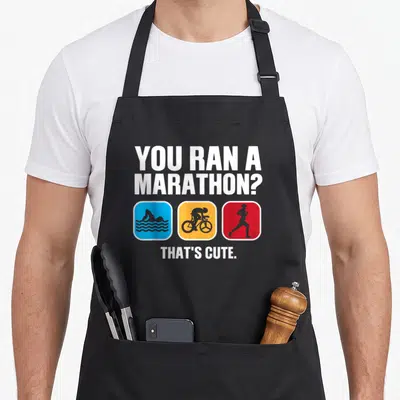 Swim run bike apron - YOU RAN A MARATHON THAT'S CUTE text with blue orange red swim bike run icons — shown on a black cotton canvas apron against a white background