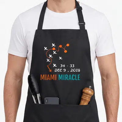 Miami comeback apron - Play diagram with 34-33 score and MIAMI MIRACLE text in orange teal white — shown on a black cotton canvas apron against a white background