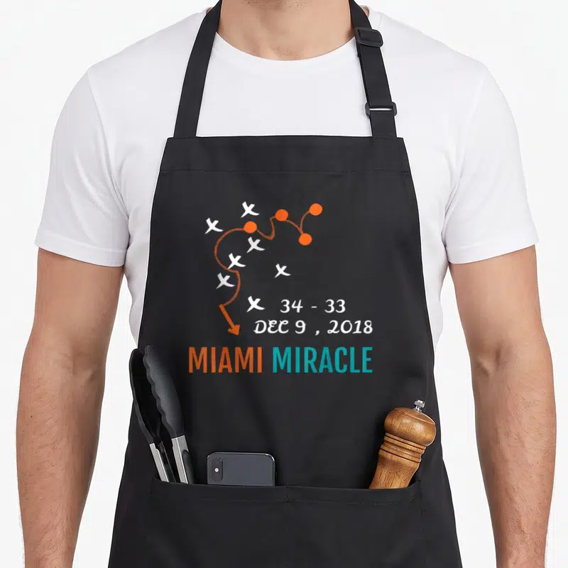 Miami comeback apron - Play diagram with 34-33 score and MIAMI MIRACLE text in orange teal white — shown on a black cotton canvas apron against a white background
