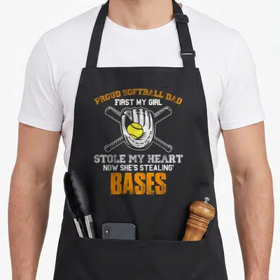 Proud softball dad apron - Softball glove holding yellow ball over two crossed bats with distressed orange and white — shown on a black cotton canvas apron against a white background