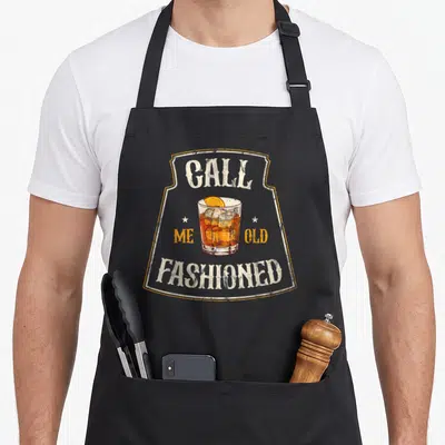 Whiskey cocktail old fashioned aprons - Retro cocktail glass with CALL ME OLD FASHIONED text and orange slice garnish — shown on a black cotton canvas apron against a white background