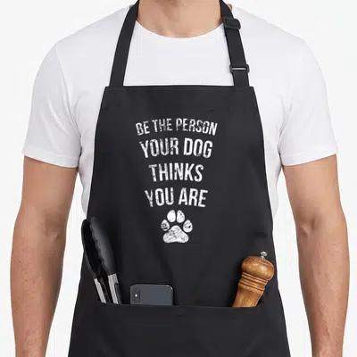 Retro pet funny dog apron - White distressed text BE THE PERSON YOUR DOG THINKS YOU ARE above a single paw print — shown on a black cotton canvas apron against a white background