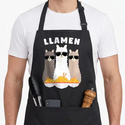 Llamen kawaii aprons - Three cartoon llamas in sunglasses holding ramen bowls under arched LLAMEN text — shown on a black cotton canvas apron against a white background