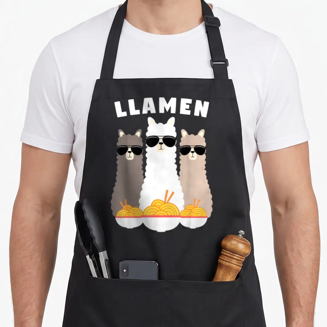 Llamen kawaii aprons - Three cartoon llamas in sunglasses holding ramen bowls under arched LLAMEN text — shown on a black cotton canvas apron against a white background