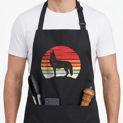 Catch coyote apron - Grey wolf silhouette standing before a retro sun with horizontal red orange yellow and blue stripes — shown on a black cotton canvas apron against a white background