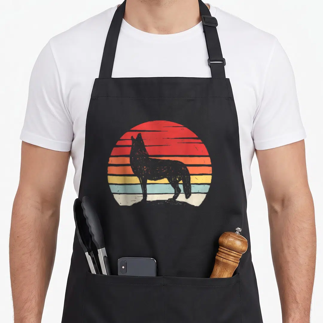 Catch coyote apron - Grey wolf silhouette standing before a retro sun with horizontal red orange yellow and blue stripes — shown on a black cotton canvas apron against a white background