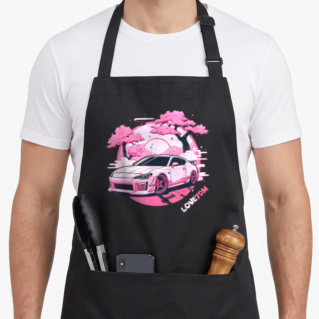 Jdm racer car apron - Retro pink car illustration with cherry trees and LOVEJDM text — shown on a black cotton canvas apron against a white background