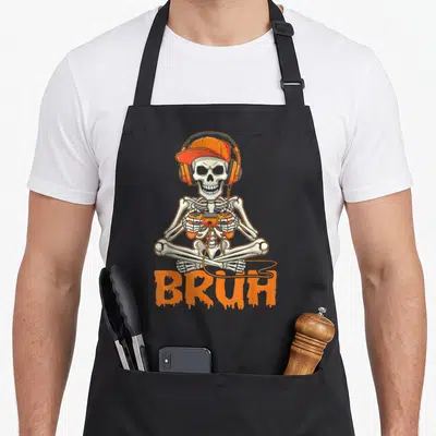 Bruh gamer halloween apron - Cartoon skeleton in orange cap and headphones holding controller above dripping BRUH text — shown on a black cotton canvas apron against a white background