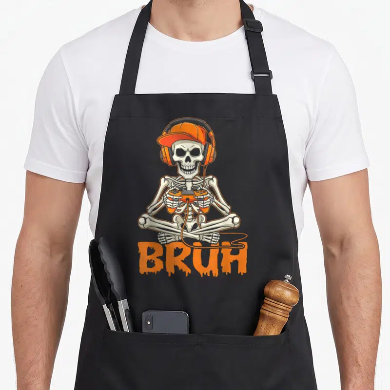 Bruh gamer halloween apron - Cartoon skeleton in orange cap and headphones holding controller above dripping BRUH text — shown on a black cotton canvas apron against a white background