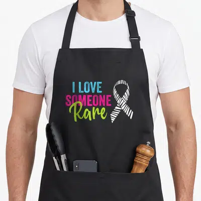 Disease zebra apron - I Love Someone Rare text in blue pink and lime green beside white striped ribbon on black — shown on a black cotton canvas apron against a white background