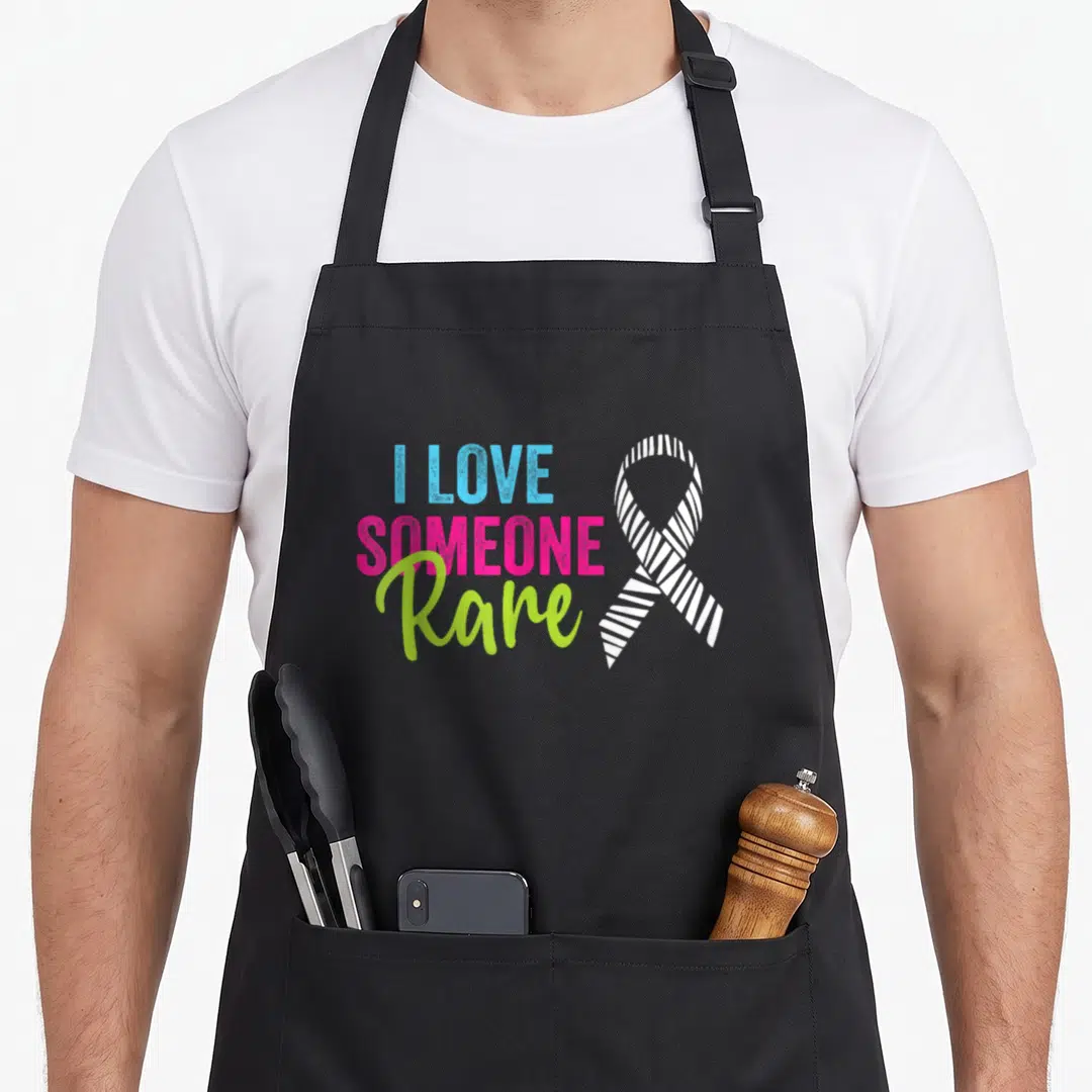 Disease zebra apron - I Love Someone Rare text in blue pink and lime green beside white striped ribbon on black — shown on a black cotton canvas apron against a white background