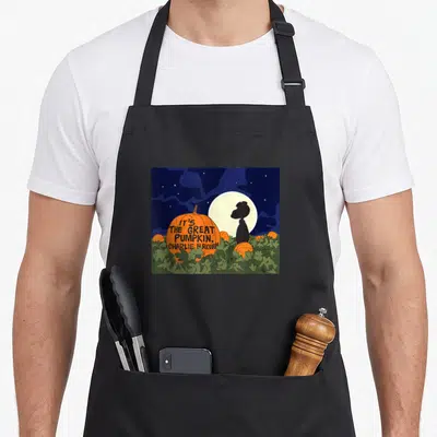 Cute pumpkin apron - Orange pumpkin with title text and character silhouette against moon in cartoon art — shown on a black cotton canvas apron against a white background