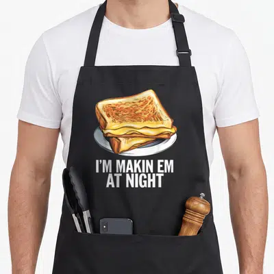 Night grilled apron - I'M MAKIN EM, AT NIGHT text above stacked golden brown grilled cheese on white plate — shown on a black cotton canvas apron against a white background