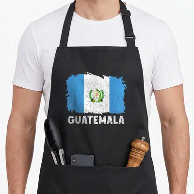 Guatemala root guatemalan aprons - Distressed blue white and green flag graphic above white GUATEMALA text — shown on a black cotton canvas apron against a white background