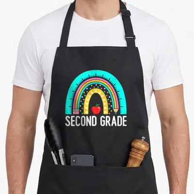 Grade rainbow 2nd apron - SECOND GRADE text under rainbow, red apple, ruler, pencil in cyan pink yellow red white — shown on a black cotton canvas apron against a white background