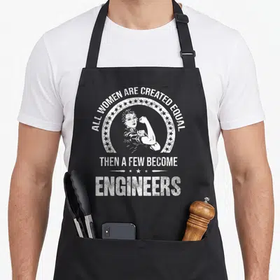 Crew engineer apron - White distressed graphic of a woman flexing arm inside star circle with engineering quote — shown on a black cotton canvas apron against a white background