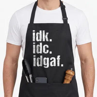 Idk birthday apron - White stacked text reading idk. idc. idgaf. in distressed font — shown on a black cotton canvas apron against a white background