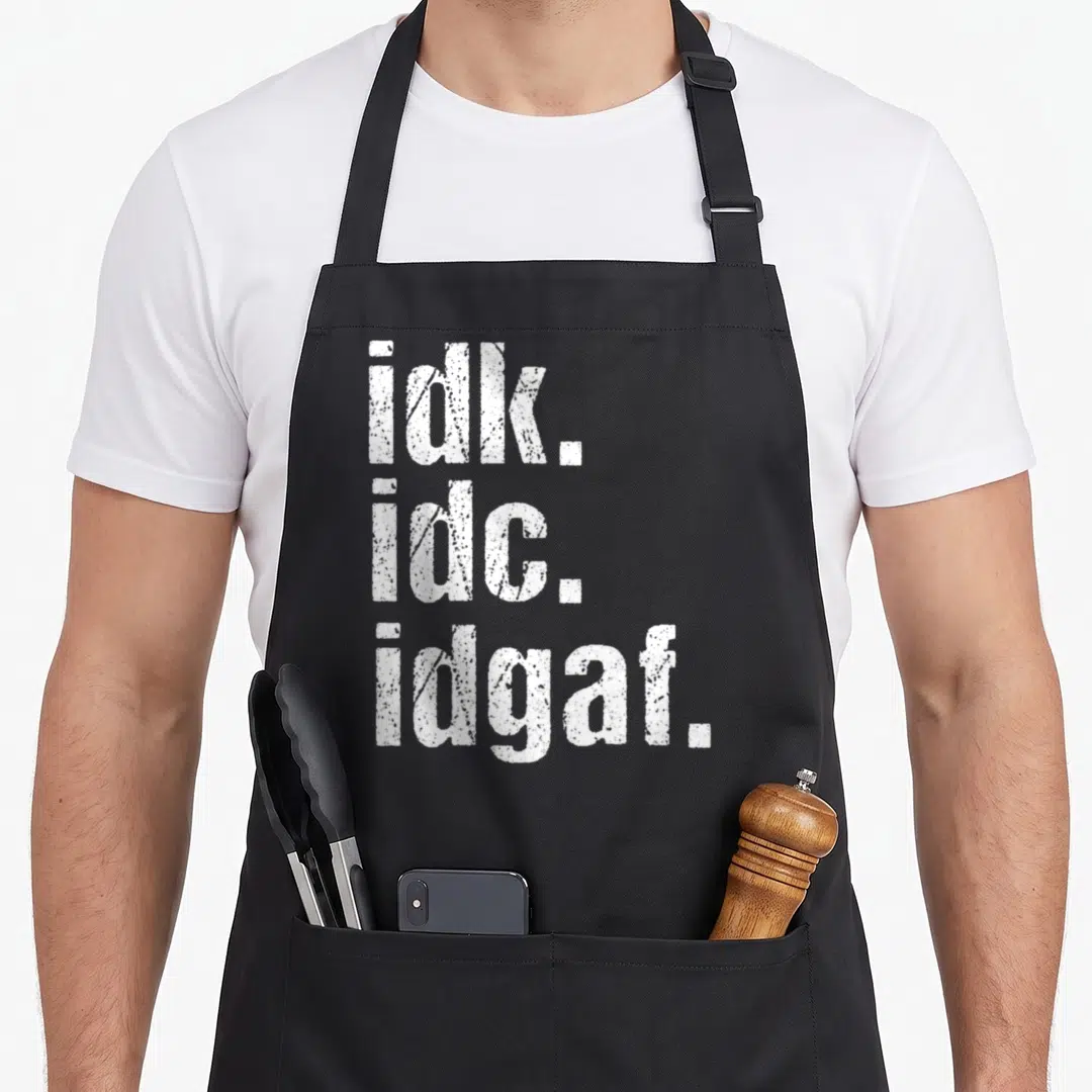Idk birthday apron - White stacked text reading idk. idc. idgaf. in distressed font — shown on a black cotton canvas apron against a white background