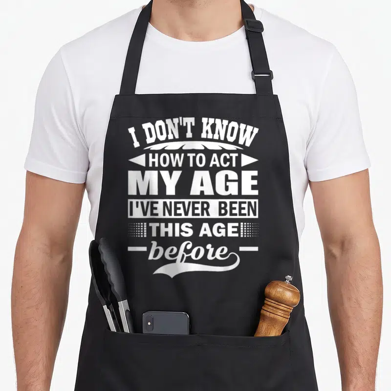 Act i love ultimate apron - White stacked text reading I DON'T KNOW HOW TO ACT MY AGE with decorative dots and lines — shown on a black cotton canvas apron against a white background
