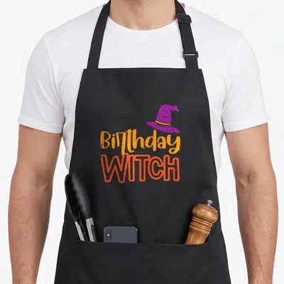 Witch funny halloween apron - Purple witch hat above stacked Birthday WITCH text in orange and red-orange letters — shown on a black cotton canvas apron against a white background