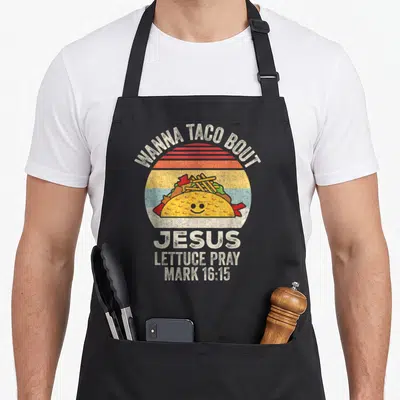 Bout christian apron - Retro smiling taco on striped sun with WANNA TACO BOUT JESUS LETTUCE PRAY text — shown on a black cotton canvas apron against a white background