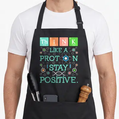 Stay chemistry apron - Periodic table tiles spelling THINK with LIKE A PROTON STAY POSITIVE text and science icons — shown on a black cotton canvas apron against a white background