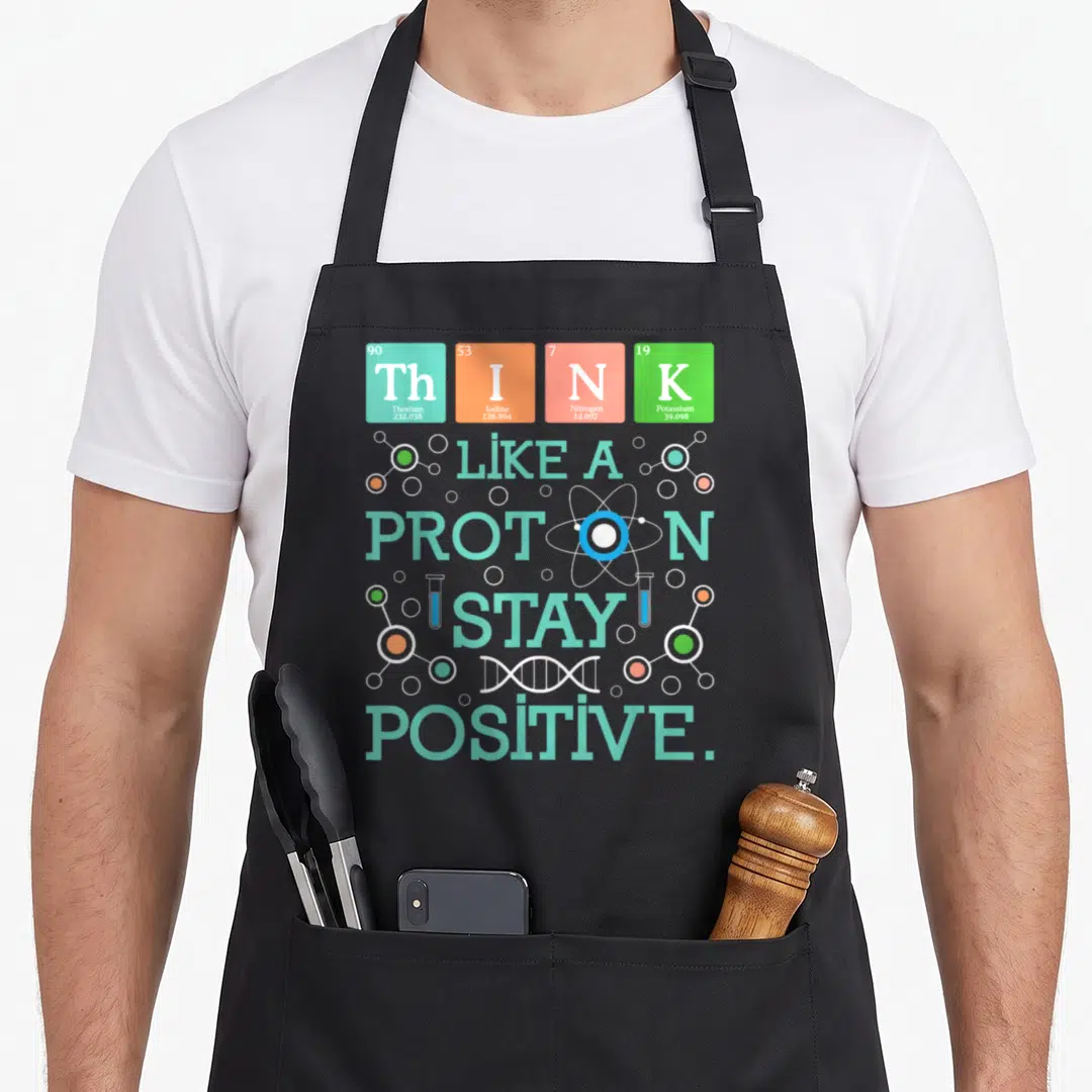 Stay chemistry apron - Periodic table tiles spelling THINK with LIKE A PROTON STAY POSITIVE text and science icons — shown on a black cotton canvas apron against a white background