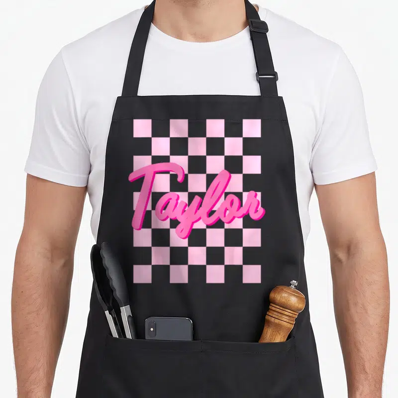 Taylor birthday apron - Pink 3D script Taylor text over grey and pink checkerboard pattern — shown on a black cotton canvas apron against a white background