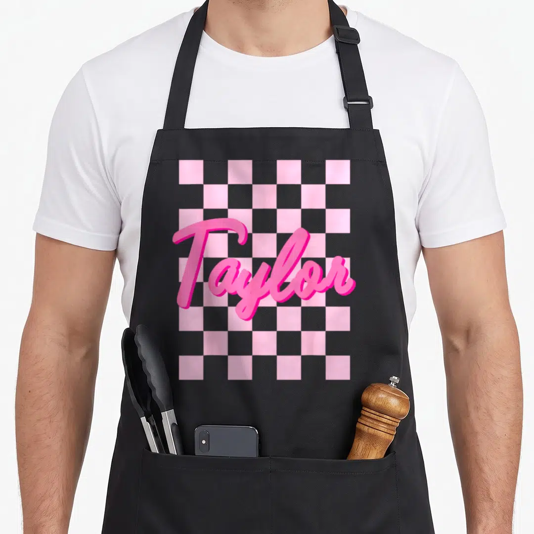 Taylor birthday apron - Pink 3D script Taylor text over grey and pink checkerboard pattern — shown on a black cotton canvas apron against a white background