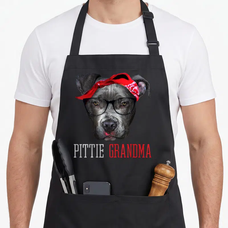 Pittie granny apron - Grey dog head wearing black glasses and red bandana with PITTIE GRANDMA text — shown on a black cotton canvas apron against a white background