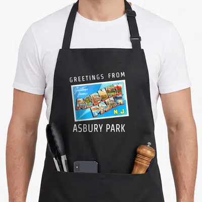 Asbury travel aprons - Retro postcard reading Greetings from Asbury Park N.J. with 3D scene-filled letters on blue — shown on a black cotton canvas apron against a white background