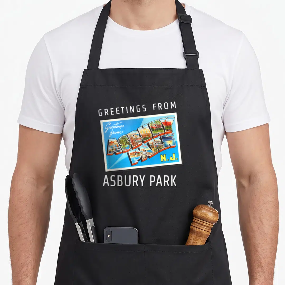 Asbury travel aprons - Retro postcard reading Greetings from Asbury Park N.J. with 3D scene-filled letters on blue — shown on a black cotton canvas apron against a white background